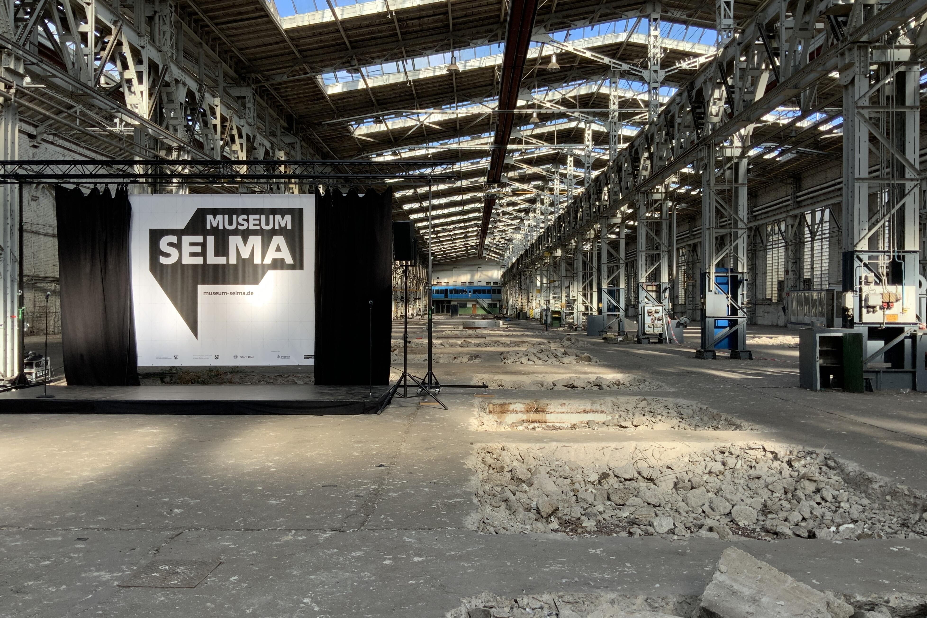 Auf dem Bild ist eine alte Industriehalle zu sehen mit aufgerissenem Betonboden und hellem Lichteinfall aus den Oberlichtern des Hallendachs. Am linken Bildrand ist eine Bühne aufgebaut und ein schwarzer Vorhang geöffnet. Dahinter ist das Logo von Museum Selma zu sehen.