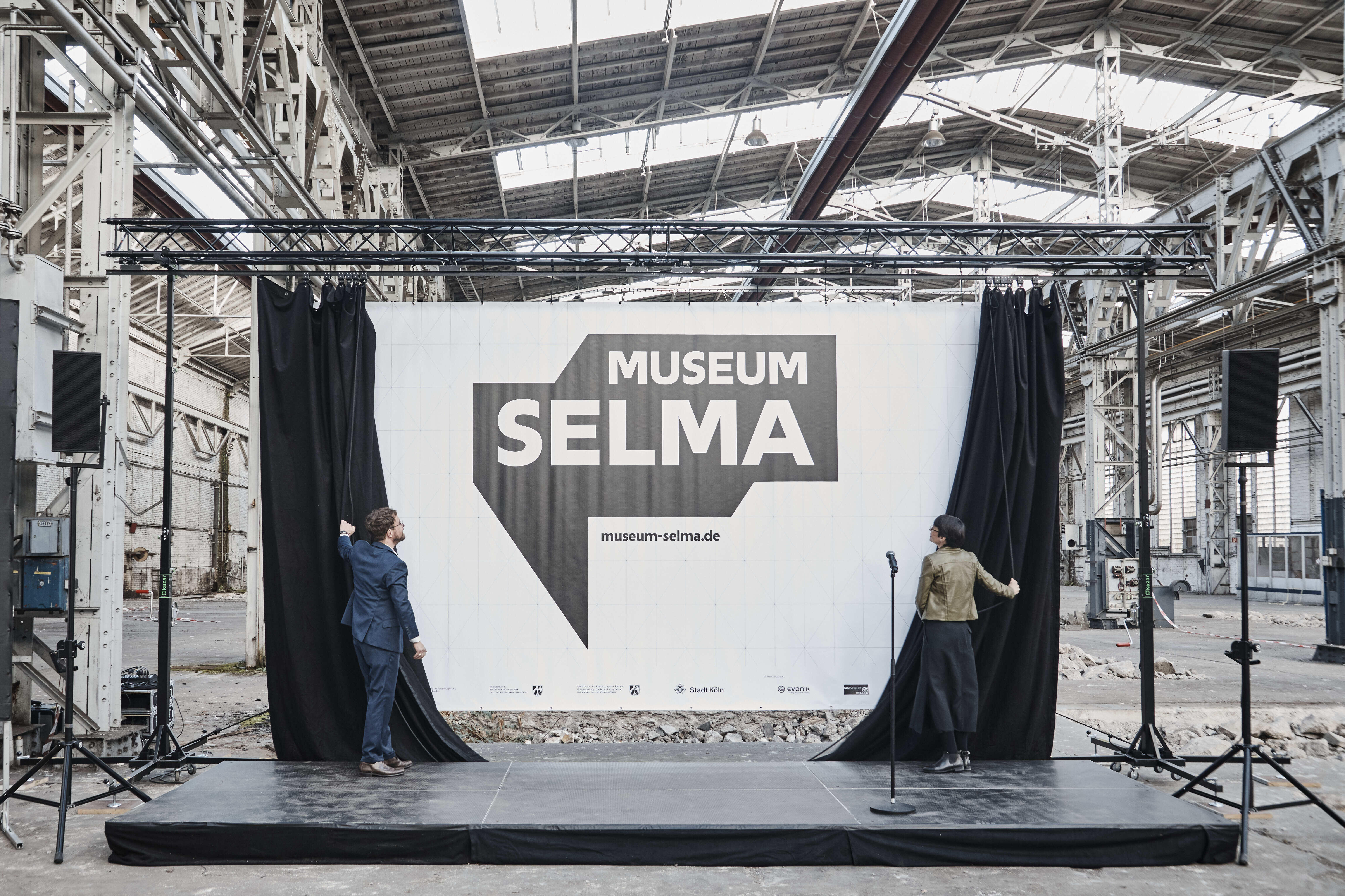 DOMiD-Geschäftsführer Dr. Robert Fuchs und NRW-Integrationsministerin Josefine Paul enthüllen den Vorhang des neuen Logos von Museum Selma. Im Hintergrund ist die alte Industriehalle zu sehen, in der das Museum entsteht.