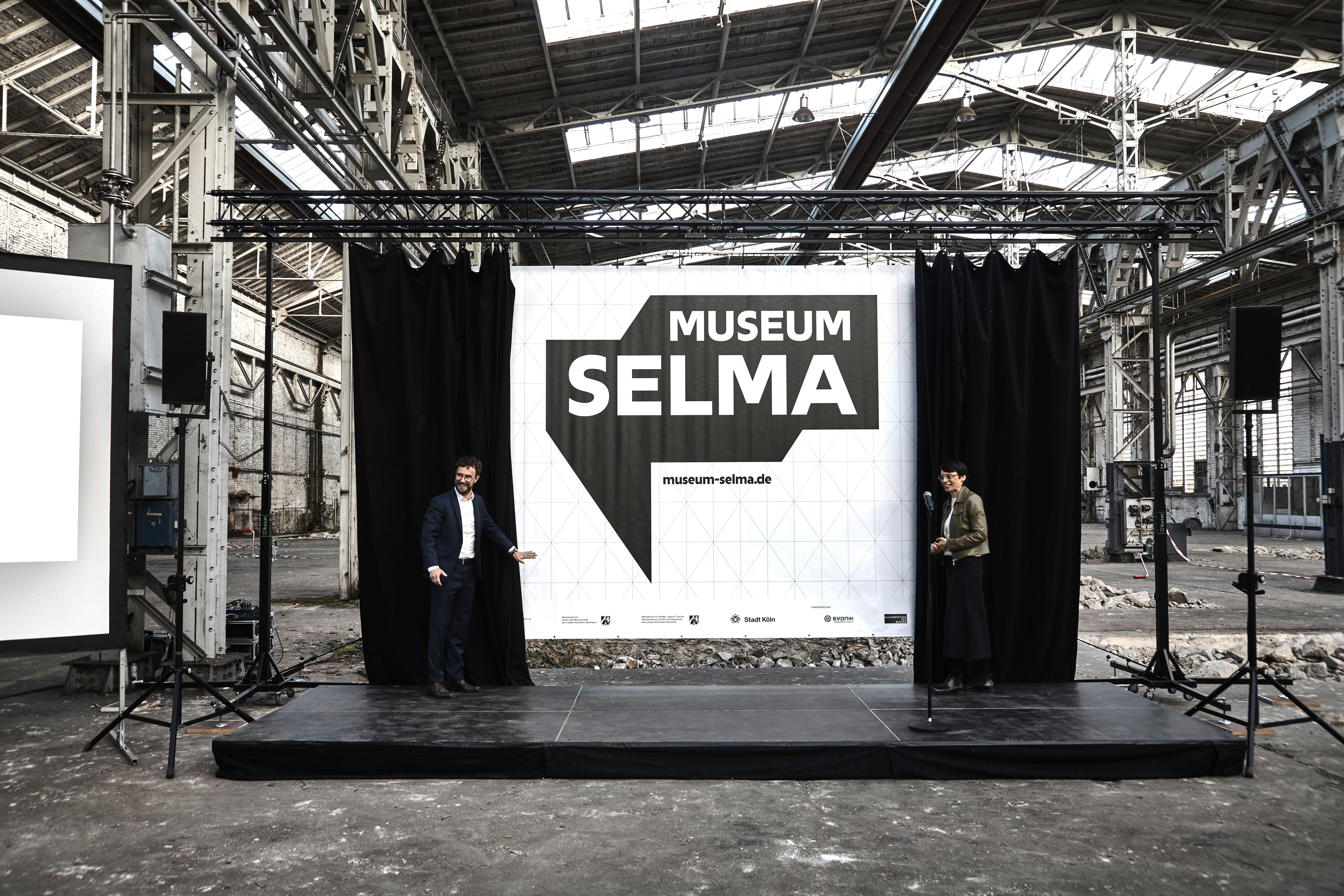 DOMiD-Geschäftsführer Dr. Robert Fuchs und NRW-Integrationsministerin Josefine Paul auf der Bühne vor dem neuen Logo von Museum Selma.