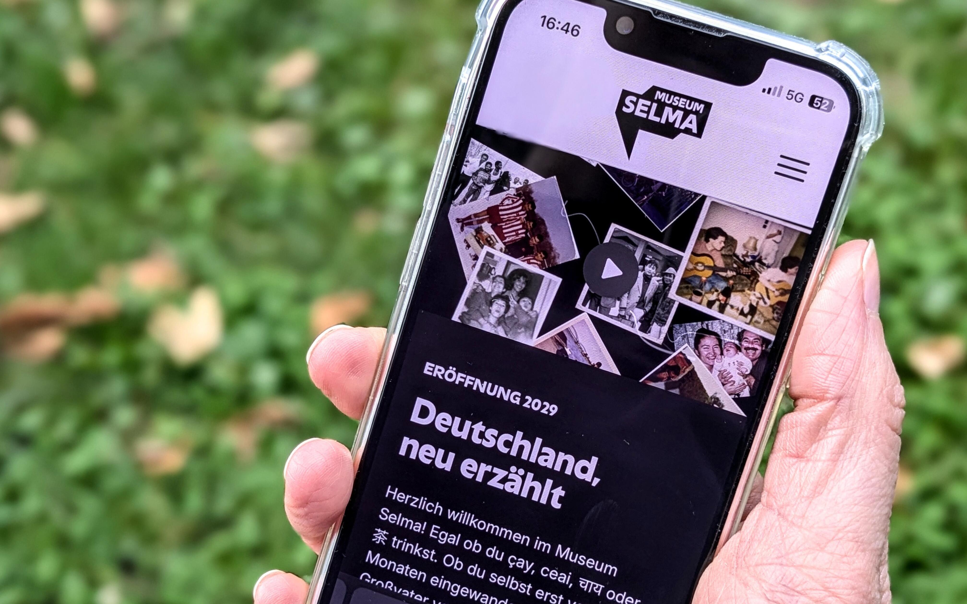 Eine Hand hält ein Smartphone, das den Startbildschirm der Museum Selma Webseite zeigt.