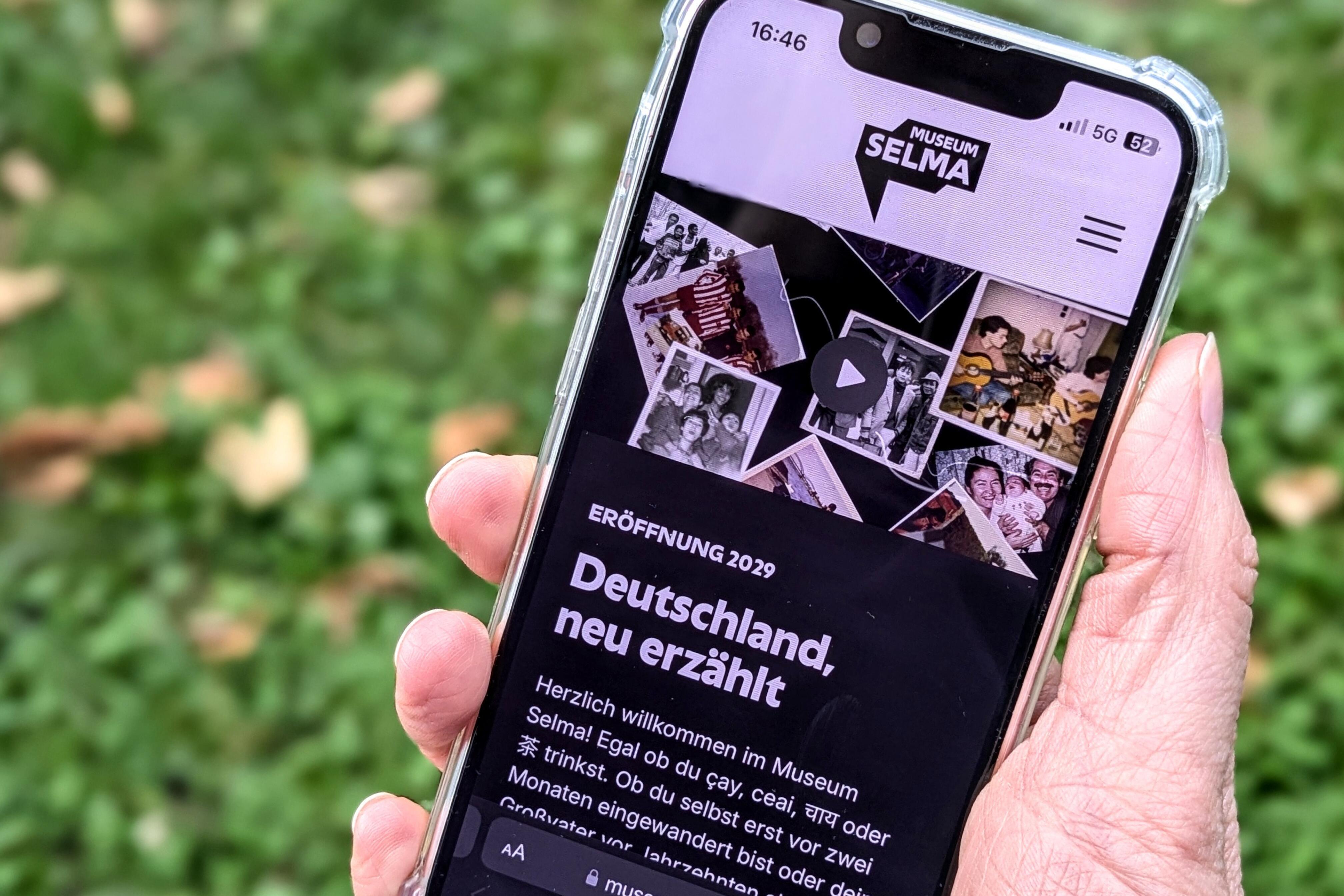 Eine Hand hält ein Smartphone, das den Startbildschirm der Museum Selma Webseite zeigt.