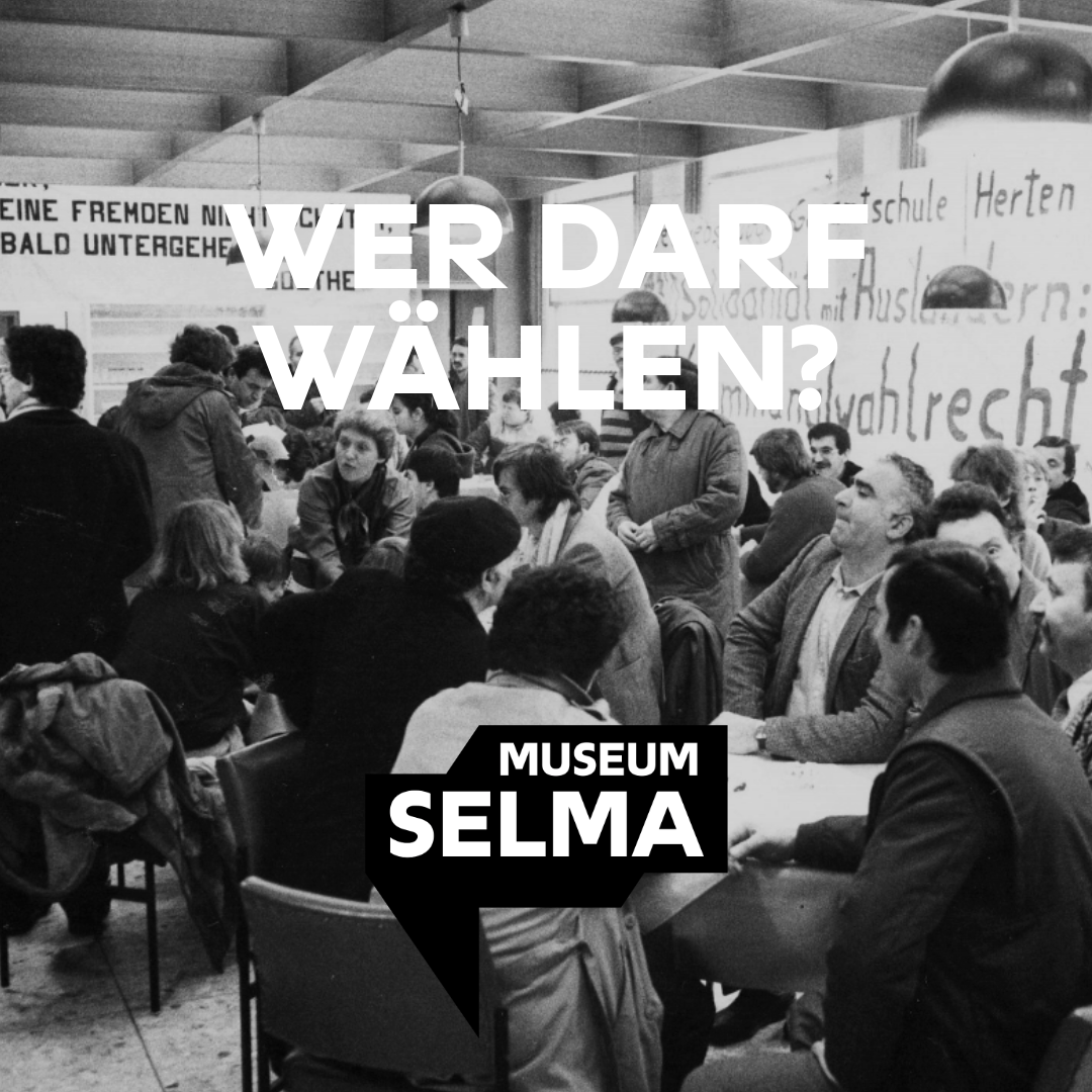 Das Foto zeigt eine Aktion zum Ausländerwahlrecht in Hessen in den 1980er Jahren. Eine Gruppe Menschen sitzt zusammen an Tischen vor Transparenten.